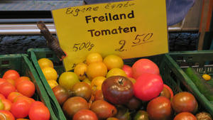 Im Mittelpunkt steht eine grne Kiste eines Verkaufsstandes auf einem Markt, die verschiedenfarbige Tomaten enthlt. An die Kiste ist mit einer Klammer eine gelbes Schild geheftet und besagt: "Eigene Ernte. Freiland Tomaten. 500 Gramm. 2,50 Euro.