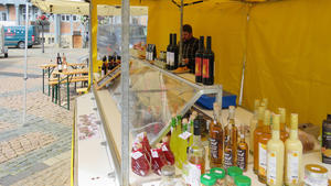 Hinter einem Verkaufsstand auf dem Wolfenbtteler Wochenmarkt steht ein Mann. Auf Tischen und in der Auslage stehen verschiedene Flaschen Wein, Essig und le. Auch weitere Lebensmittel sind zu erkennen.
