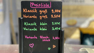 Auf einem schwarzen Preisaufsteller sind in verschiedenen Neonfarben fnf verschiede Cheesecake-Varianten und zugehrige Preise ausgewiesen.