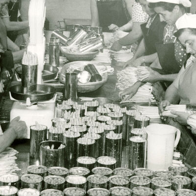 Schwarzweifoto: Frauen in Kittel und mit Kopfhaben stehen an einem tisch und bereiten Spargel vor bzw. stecken ihn in Konservendosen